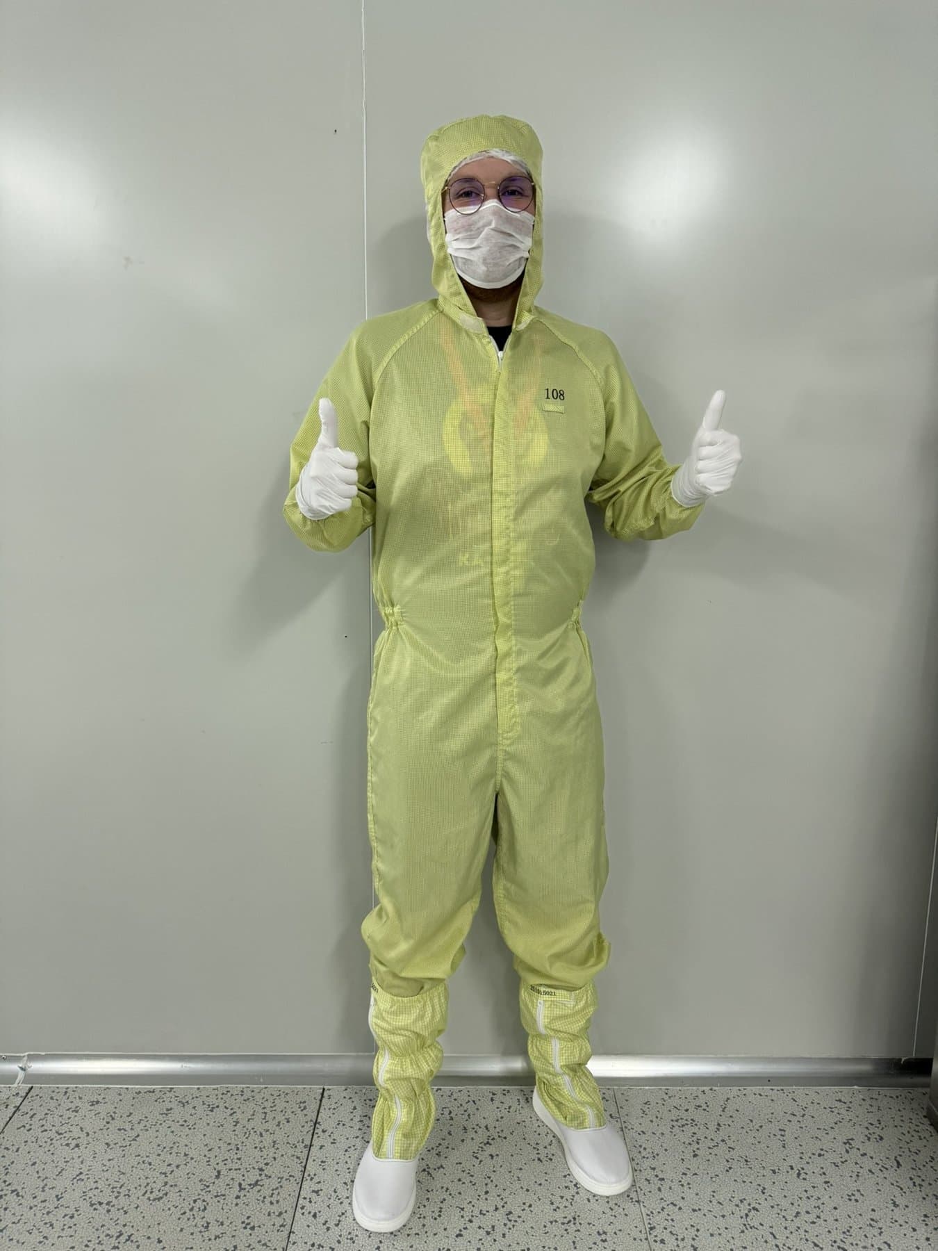 Frank in a pale green cleanroom coverall, hood, mask and gloves, giving two thumbs up against a plain grey wall