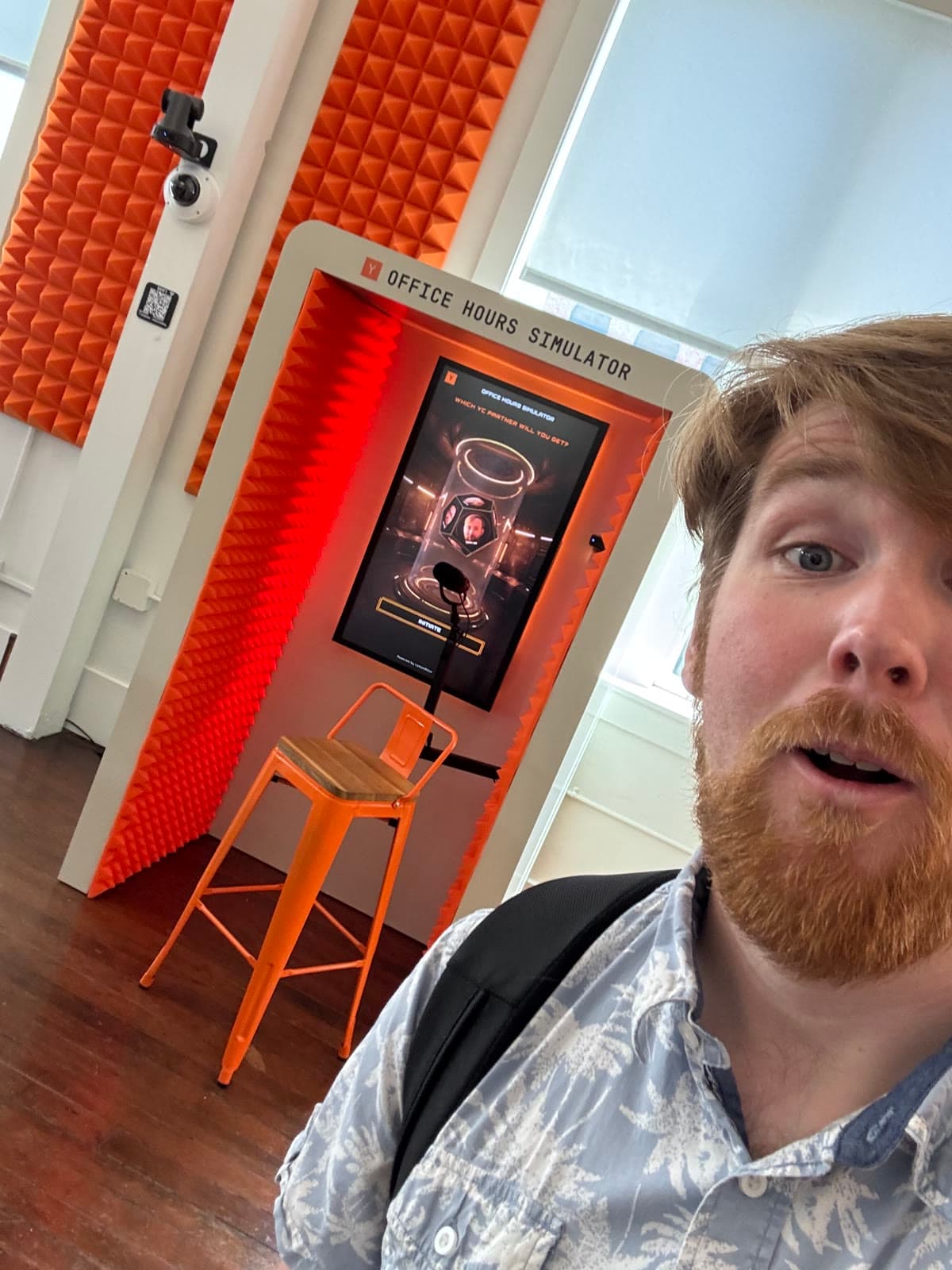 Selfie with the YC Office Hours Simulator booth: an orange acoustic-foam booth with a mic, a stool, and a screen reading WHICH YC PARTNER WILL YOU GET?